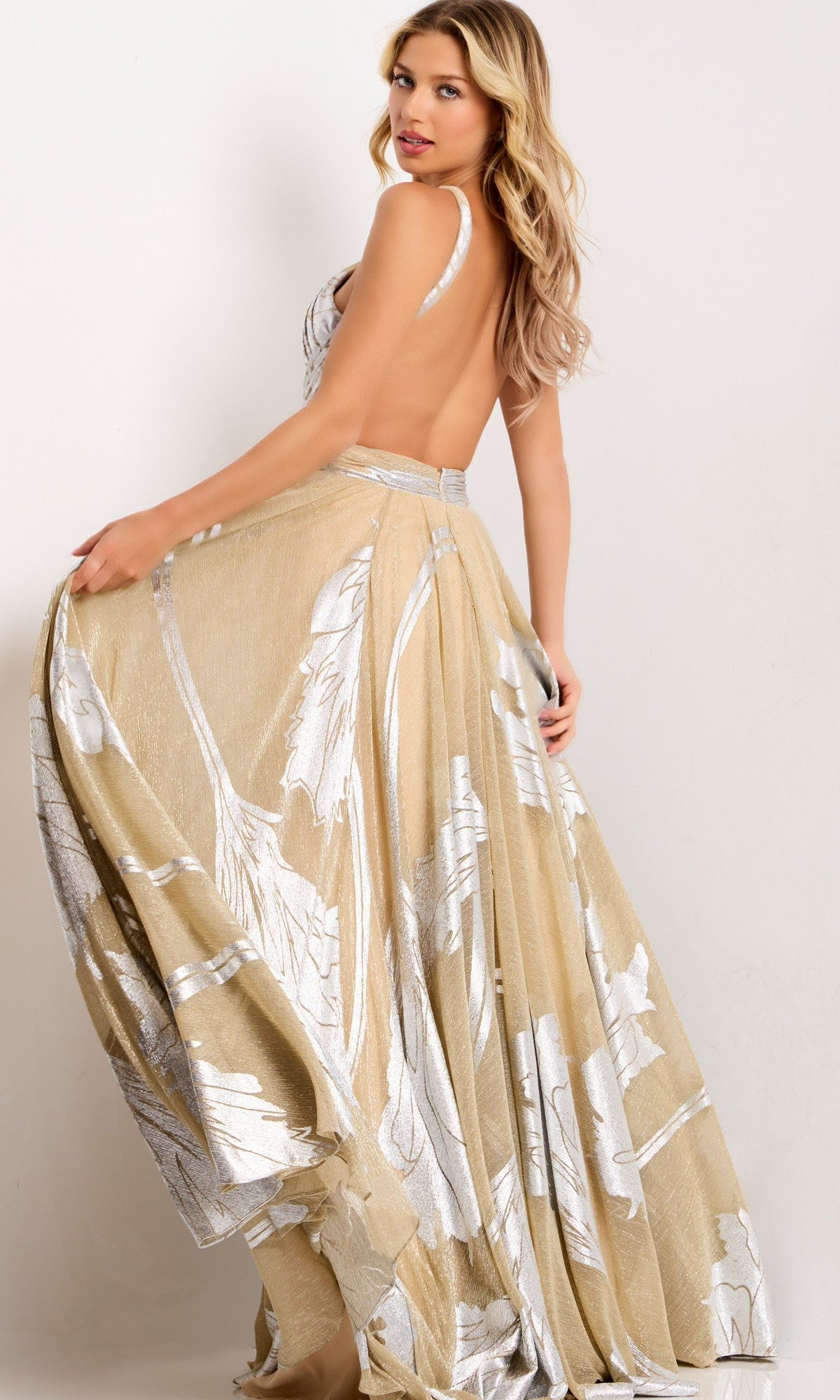 A woman poses sideways in the Jovani 44421 long prom dress—a backless gold and nude gown with silver metallic leaf accents—holding the flowing skirt and glancing over her shoulder against a simple light background.